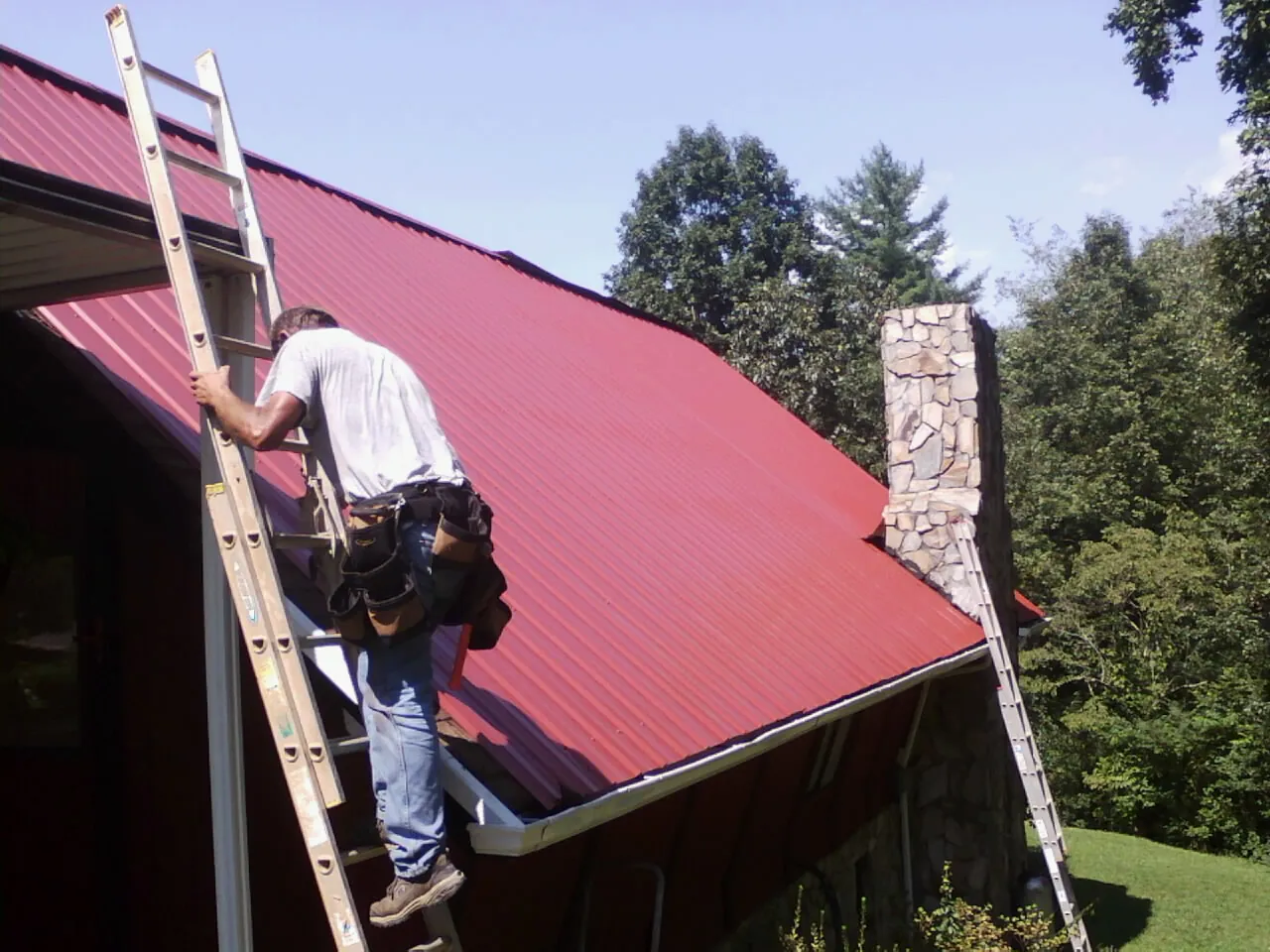Roofer on a ladder beside a red metal roof and stone chimney — Soffit & Fascia Repair by Roofing Brothers in Three Forks